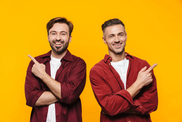Image of two handsome men 30s in red shirts smiling and pointing fingers upward at copyspace isolated over yellow background - Valokuva, kuva