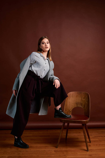 Independent young woman posing in the photostudio. Gorgeous young female in classic suit and grey coat. Brown background. Vintage chair style. Woman dressed in Man clothes. Fashionable clothes. - Foto, Imagem
