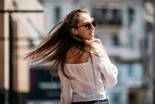 stylish smiling happy woman dressed in white shirt.young beautiful brunette walking the city. She looks back and smile.hair develops in the wind - Fotoğraf, Görsel