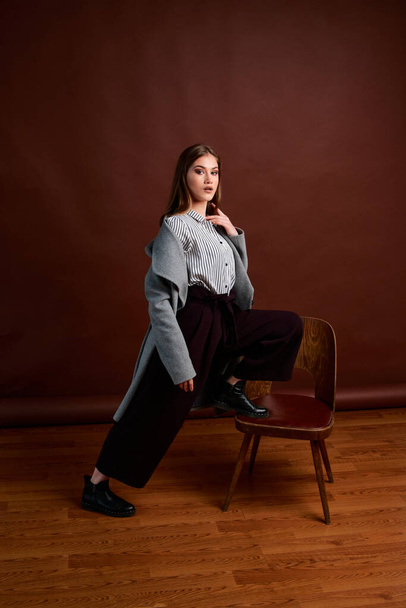 Independent young woman posing in the photostudio. Gorgeous young female in classic suit and grey coat. Brown background. Vintage chair style. Woman dressed in Man clothes. Fashionable clothes. - Foto, afbeelding