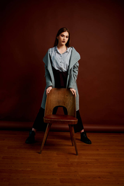 Independent young woman posing in the photostudio. Gorgeous young female in classic suit and grey coat. Brown background. Vintage chair style. Woman dressed in Man clothes. Fashionable clothes. - 写真・画像