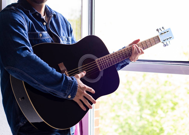 Muž v klobouku s bradkou hraje v létě na kytaru na balkóně. Boční pohled. Covid - 19 - Fotografie, Obrázek