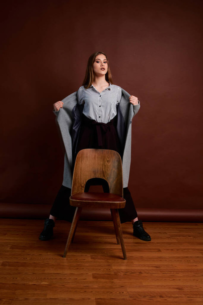 Independent young woman posing in the photostudio. Gorgeous young female in classic suit and grey coat. Brown background. Vintage chair style. Woman dressed in Man clothes. Fashionable clothes. - Foto, Imagen