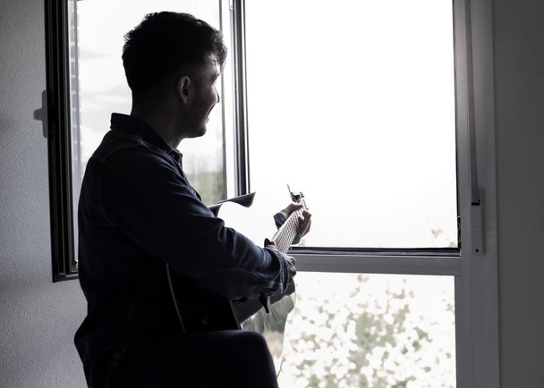 mężczyzna w domu przy oknie i gra na gitarze akustycznej - Zdjęcie, obraz