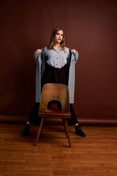 Independent young woman posing in the photostudio. Gorgeous young female in classic suit and grey coat. Brown background. Vintage chair style. Woman dressed in Man clothes. Fashionable clothes. - Photo, Image