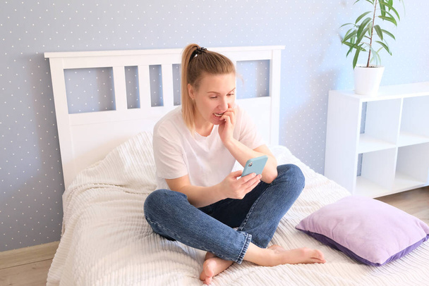 kaunis nainen poseeraa istuessaan sängyllä älypuhelin käsissään
 - Valokuva, kuva