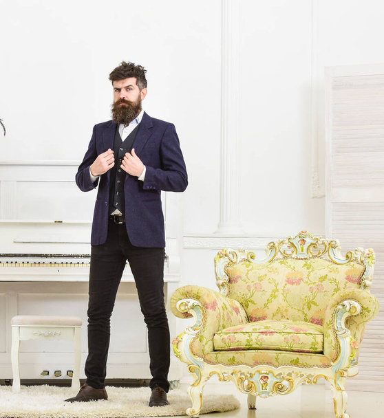 Macho attractive and elegant on serious face buttoning up his jacket. Elegant outfit concept. Man with beard and mustache wearing classic suit, stands near old fashioned armchair, white background. - Foto, immagini