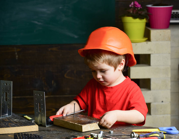 Kid boy in orange hard hat or helmet, study room background. Childhood concept. Boy play as builder or repairer, work with tools. Child dreaming about future career in architecture or building. - Foto, Bild