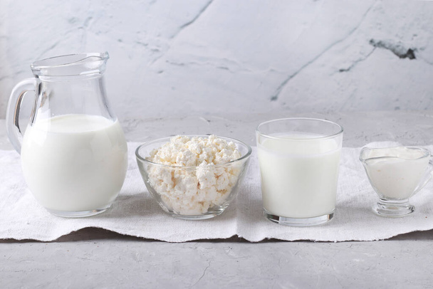 Молочные продукты: молоко, кефир или айран, творог и сметана в прозрачной миске, кувшин и стекло на сером фоне, горизонтальная ориентация
 - Фото, изображение