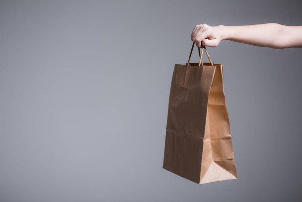 Female hand on a gray background holds out or picks up a paper bag with handles. Advertising shooting for a company with delivery. - Фото, изображение