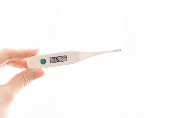 Die Finger einer weiblichen Hand halten ein elektronisches Thermometer mit einer Temperatur von 35,7 Grad auf einer Anzeigetafel auf weißem Hintergrund. Pathologie, Krankheit, Unterkühlung. - Foto, Bild