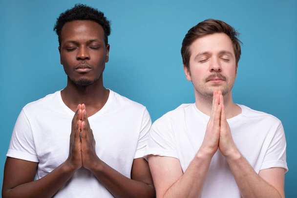 Hopeful adult men closing eyes and pressing palms together, praying, hoping for the best. Calm peaceful guys holding hands in namaste while meditating - Φωτογραφία, εικόνα