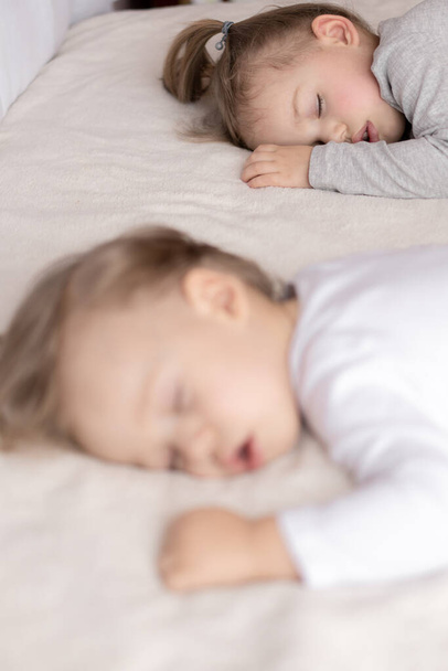 Childhood, sleep, relaxation, family, lifestyle concept - two young children 2 and 3 years old dressed in white and beige bodysuit sleep on a beige and white bed at lunch holding hands top view. - 写真・画像