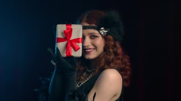 Χαρούμενη vintage styled γυναίκα ντυμένη στα 20 χρόνια Roaring κρατώντας κουτί δώρου με φιόγκο σε βελούδινο φόντο. Χαίρεται που είναι παρούσα. ρετρό, κόμμα, έννοια της μόδας. - Πλάνα, βίντεο