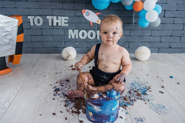 Babys first birthday - the boy sits in a space set with a robot and balloons in the background and ruins his delicious blue cake - Foto, Bild