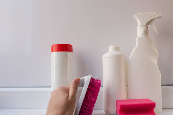 Ferramentas para limpar a casa. Gel de limpeza, pó e spray para limpeza e desinfecção de várias superfícies em recipientes brancos de plástico com uma escova na mão e uma esponja rosa sobre um fundo branco
. - Foto, Imagem