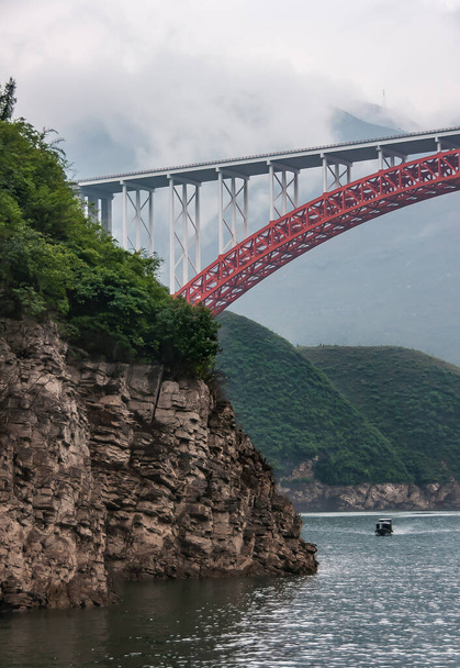Wuchan, China - 7 травня 2010: Dragon Gate Gorge on Daning River. Червоний лук з білим бетонним дорожнім мостом над темно-зеленою водою під туманним небом. Гірський берег і човен.. - Фото, зображення