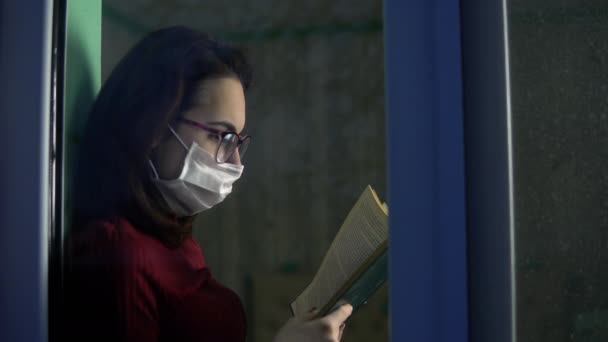 A young woman is sitting at home in quarantine with a book in her hands. A girl sits on a window sill in a mask and reads a book. View behind the glass. - Metraje, vídeo