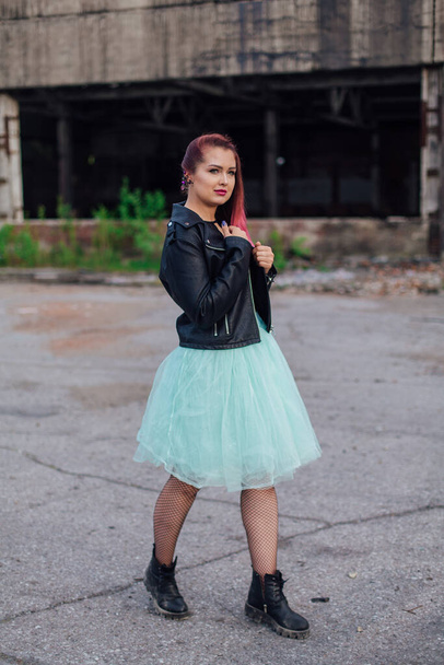 Portrait of a young girl with pink hair dressed in leather jacket and a blue skirt standing outside of collapsed building - Zdjęcie, obraz