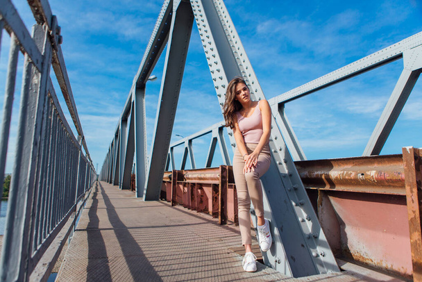 Warm summer portrait of a young beautiful woman posing on the old rusty transport bridge over the river during sunset. - Zdjęcie, obraz