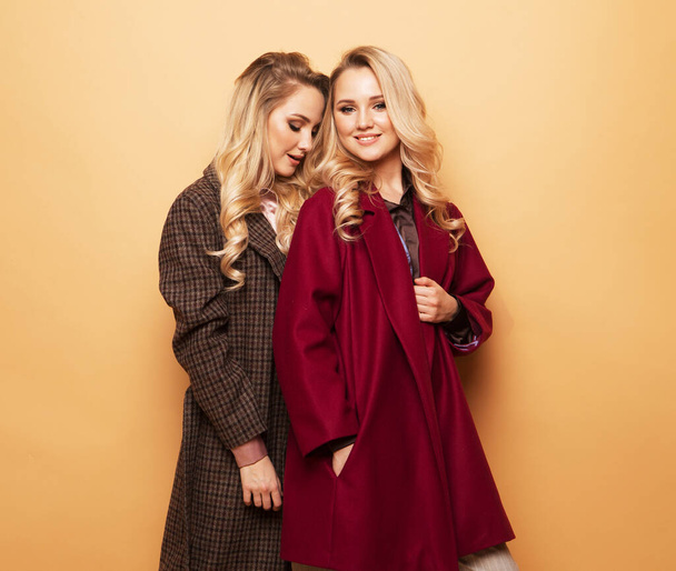portrait of two girls, best friends posing indoor on beige background wearing winter stylish coat. - Fotó, kép