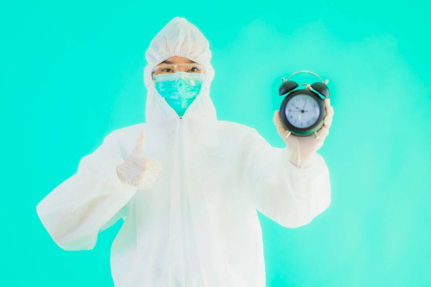Портрет красивая молодая азиатская врач женщина носить ppe Suite и маска показать часы или будильник на голубом изолированном фоне
 - Фото, изображение