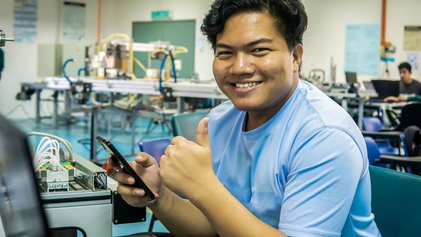 Een jonge ingenieursstudent uit Maleisië die een smartphone vasthoudt en duimen laat zien met het automatiseringssysteem als achtergrond in het laboratorium. Afbeelding bevat geluidsreductie. Geselecteerde focus. - Foto, afbeelding