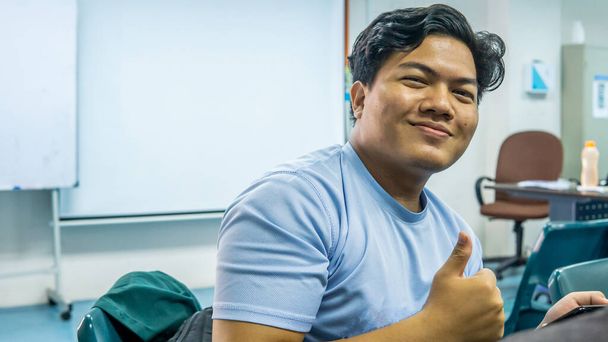 Jonge Maleise student glimlach helder en toont een duim omhoog of een goed teken in de klas. Het gebruik van een smartphone of gadget voor onderwijs. - Foto, afbeelding