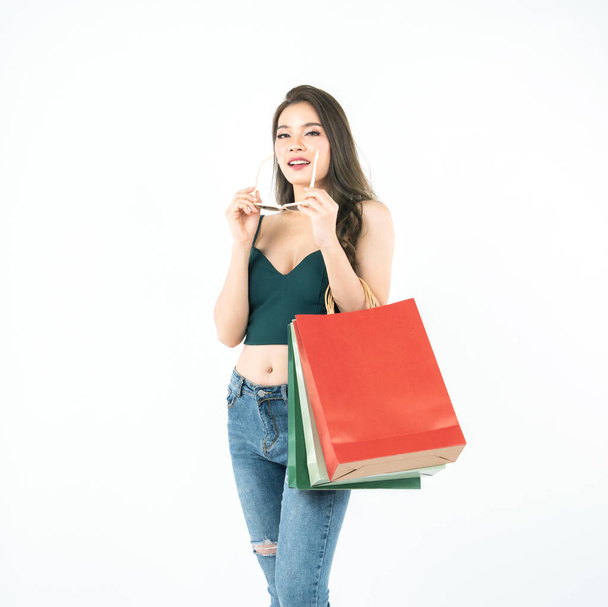 Gelukkig jong aziatisch vrouw in groen sexy shirt en jean geniet van winkelen, hand houden zonnebril en kleurrijke boodschappentassen, verkoop opgewonden, lente zomer mode trend geïsoleerd op witte achtergrond. - Foto, afbeelding
