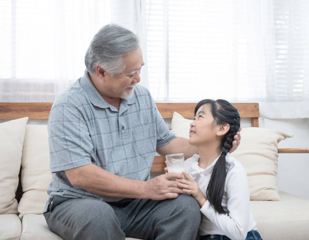 feliz asiático mayor anciano abuelo tener nieto cuidar y cuidar con dar leche y beso en mejilla mientras sentado en sofá en casa, retiro salud estilo de vida concepto
. - Foto, imagen