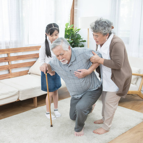 Aziatische grootvader vallen grootmoeder en kleindochter hulp en ondersteuning dragen hem om te zitten op de bank, pensioen leven concept. - Foto, afbeelding