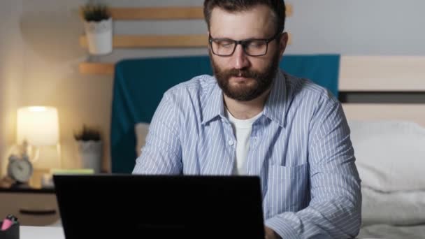 A fickó otthon dolgozik a számítógépnél. A szakállas fickó asztalhoz ül a hálószobában, és gépel a laptop billentyűzetén. Otthoni munka, szabadúszó koncepció - Felvétel, videó