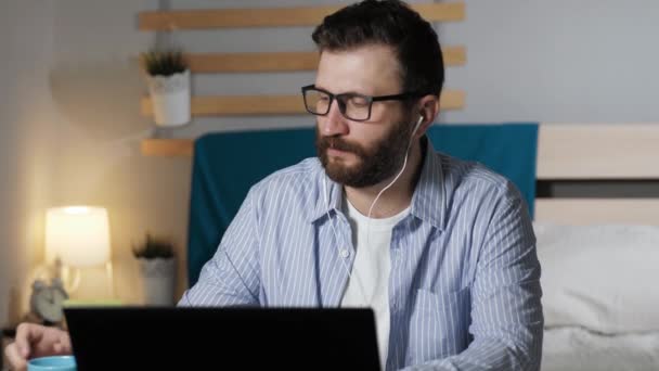 Man drinks coffee listens to music and works at home. Bearded guy in headphones sits at table in bedroom, works on laptop, drinks coffee or tea and listens to music. Work at home, freelance concept - Video, Çekim