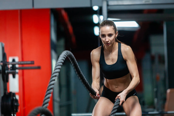 An attractive young muscular girl using training ropes for battle ropes exercise in a gym - Foto, Imagem