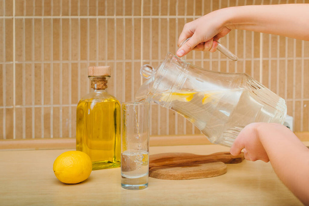 Dívka nalévá vodu s citrónem ze džbánu do sklenice v domastické kuchyni. Koncept zdravého života - Fotografie, Obrázek
