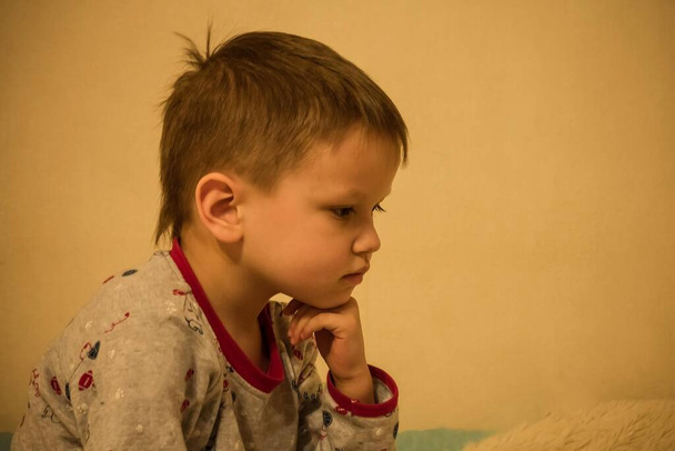 The little boy propped up his chin with his fist. Face in profile, look down. Thoughtfulness, concentration. Light background, home furnishings, copy space. - Photo, Image