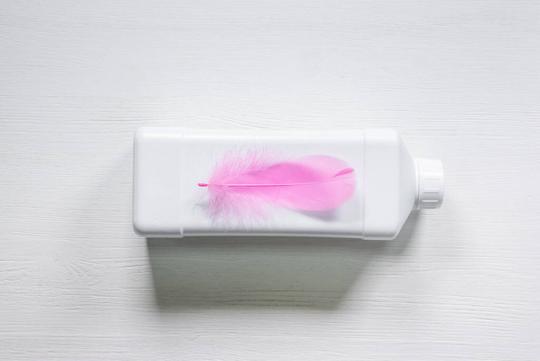 White detergent bottle and pink bird feather on white wooden table flat lay background. - Фото, изображение