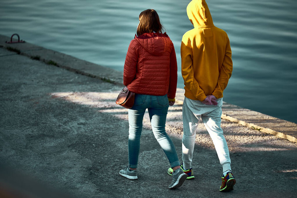 Egy sárga kabátos fickó és egy piros kabátos lány sétálnak a kikötőben a víz mellett, és elöl néznek. Csend, béke és természet. létra a vízhez. utcai fotó. - Fotó, kép