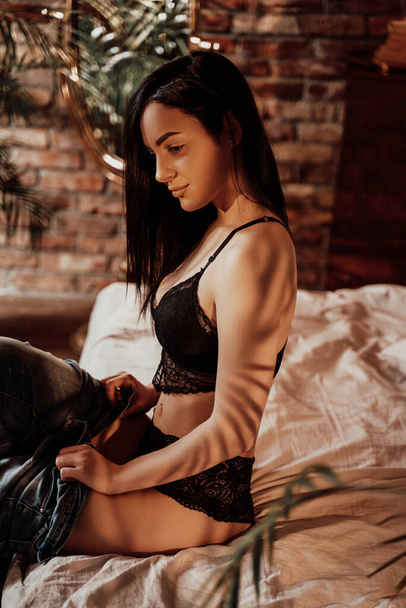 Sensual young woman sitting on the bed in fron of the mirror in a luxury looking flat, wearing hot lace underwear and taking her jeans off - Fotó, kép