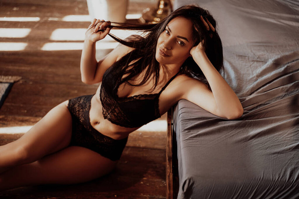 Close-up portrait of a young caucasian woman sittin on the wooden floor and leaning on the bed, wearing sexy lace lingerie and looking relaxed and pleased - Foto, Bild