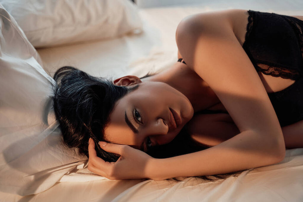 Primo piano di una splendida e sexy modella con i capelli castani che indossa lingerie di pizzo mentre posa in un letto accogliente alla luce del sole
 - Foto, immagini