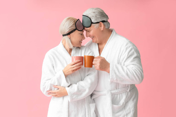 volwassen paar in badjassen en met slaap maskers drinken thee op kleur achtergrond - Foto, afbeelding