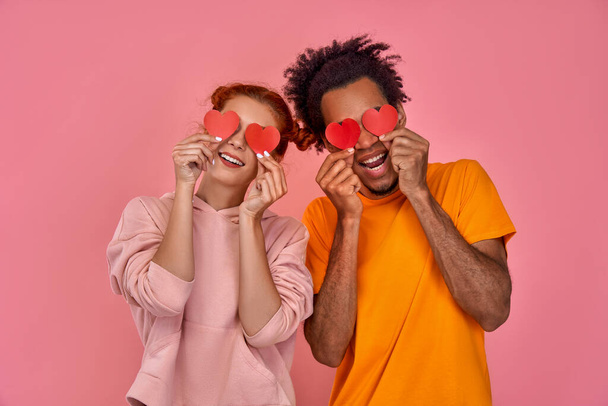 Handsome African American man and red haired caucasian woman blinded by great love for each other. Young cheerful mixed race couple in love holding red hearts over eyes and smiling, pink wall - Foto, afbeelding