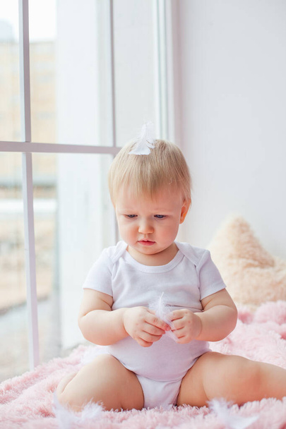 Uma menina caucasiana pequena senta-se sozinha no peitoril da janela e brinca com penas brancas
. - Foto, Imagem