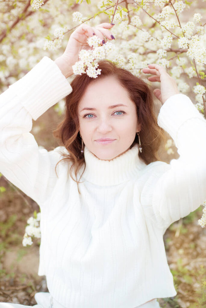 Pretty woman with brown hair in a white knitted sweater in spring in a blooming cherry garden. Nature in spring - Фото, изображение