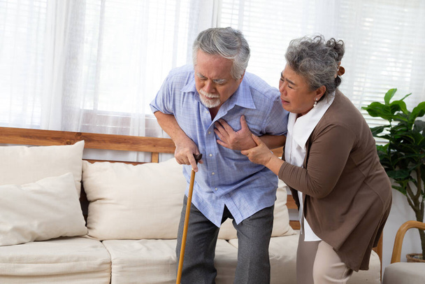 Ασιάτης ηλικιωμένος άνδρας με πόνο στην καρδιά στο σαλόνι και να φροντίσει από τη σύζυγό του, πρόβλημα υγείας και αίσθημα ασθένειας - Φωτογραφία, εικόνα