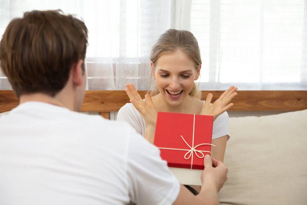 Hombre joven dando un regalo caja regalo a la novia hermosa mujer en el sofá en casa en la sala de estar, esposa sorprendida emocionada y sintiéndose sorprendida por el marido, día de San Valentín, las relaciones de pareja
 - Foto, imagen