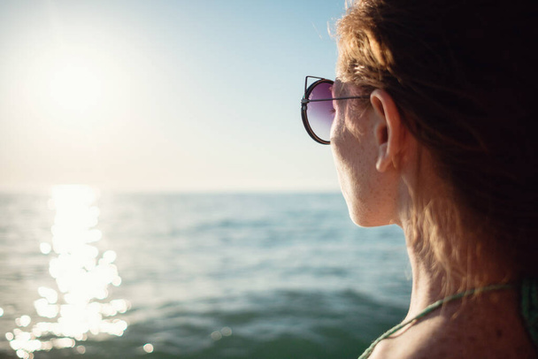 Ingwermädchen mit Sommersprossen im Bikini blickt auf einen wunderschönen Sonnenuntergang über dem Meer. Idee und Konzept von Entspannung, Freiheit, Urlaub und Glück - Foto, Bild