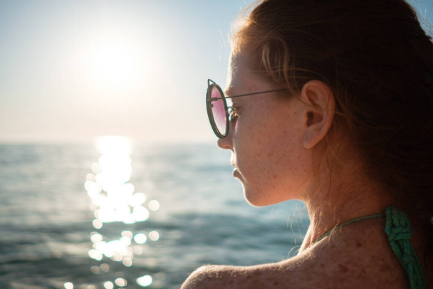 Chica pelirroja con pecas en bikini mira una hermosa puesta de sol sobre el mar. Idea y concepto de relajación, libertad, vacaciones y felicidad
 - Foto, Imagen