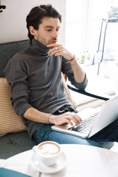 Image of handsome caucasian young man working on laptop computer and drinking coffee in modern cafe - Фото, зображення